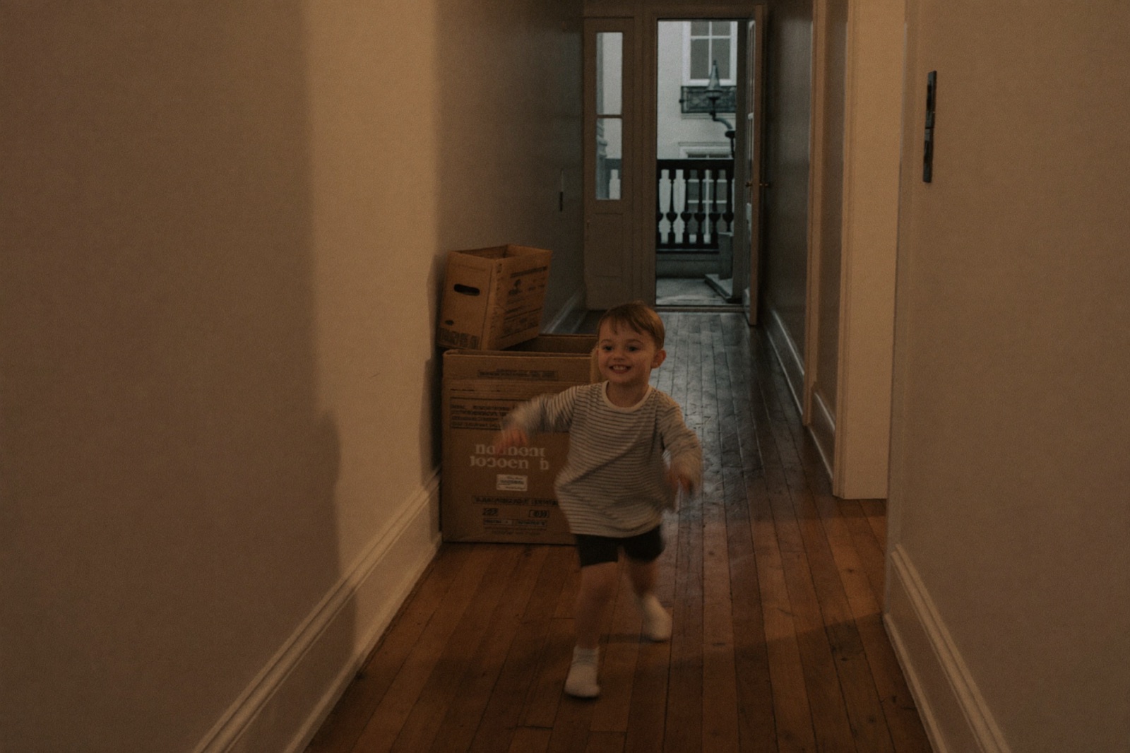 A young child running down the bare hallway of a just-purchased Victorian terrace, moving boxes by the wall.